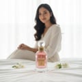 Maison Alhambra So Candid by Maison Alhambra 100 ml EDP — A pink perfume bottle with a gold cap sits on a white bedspread, with two small white flowers beside it. A woman in a white robe is blurred in the background. | Maison Alhambra So Candid by Maison Alhambra 100 ml EDP — Sebotol minyak wangi merah jambu dengan