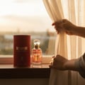 Maison Alhambra So Candid — A red cylindrical box and a clear glass perfume bottle with a gold cap and pink liquid sit on a wooden surface next to a window with sheer curtains being pulled back by hands. | Maison Alhambra So Candid — Kotak silinder merah dan botol minyak wangi kaca jernih dengan penutup emas dan
