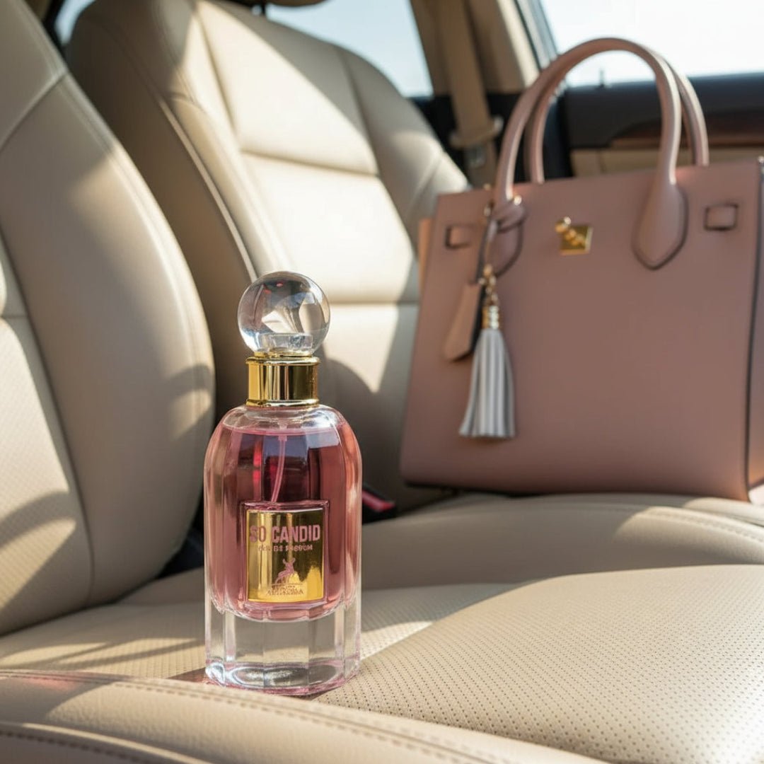 So Candid by Maison Alhambra 100 ml EDP — A pink perfume bottle with a gold label and a clear spherical cap sits on a car seat next to a blush pink handbag with a tassel. | So Candid by Maison Alhambra 100 ml EDP — Botol pewangi merah jambu dengan label emas dan penutup sfera jernih terletak di atas tempat duduk