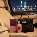 Maison Alhambra Summer Forever by Maison Alhambra 100ml EDP — A quilted black handbag and a pink glittery perfume bottle and box are placed on cream leather car seats at night with a city skyline in the background. | Maison Alhambra Summer Forever by Maison Alhambra 100ml EDP — Beg sandang kulit hitam beropol dan