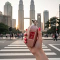 Maison Alhambra Summer Forever by Maison Alhambra 100ml EDP — A hand with bright pink nails holds a rose gold glittery perfume bottle in front of the Petronas Twin Towers in Kuala Lumpur, with a pedestrian crossing in the foreground. | Maison Alhambra Summer Forever by Maison Alhambra 100ml EDP — Tangan berkuku merah