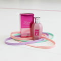 Maison Alhambra Summer Forever by Maison Alhambra 100ml EDP — Pink glitter perfume bottle and box surrounded by pastel colored elastic bands on a white textured surface. | Maison Alhambra Summer Forever by Maison Alhambra 100ml EDP — Botol dan kotak pewangi merah jambu berkilauan dikelilingi oleh gelang berwarna