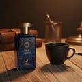Maison Alhambra True Word — A blue glass perfume bottle with a gold cap sits on a wooden surface next to a black mug and a stack of leather-bound books. | Botol kaca biru Maison Alhambra True Word dengan penutup emas terletak di atas permukaan kayu bersebelahan dengan mug hitam dan timbunan buku bersampul kulit.