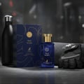 Maison Alhambra True Word by Maison Alhambra 100ml EDP — A blue perfume bottle and its box sit next to a black water bottle and a black gym glove on a grey surface, with a blurred gym background. | Maison Alhambra True Word by Maison Alhambra 100ml EDP — Sebotol minyak wangi biru dan kotaknya diletakkan di sebelah