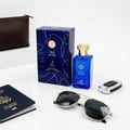 Maison Alhambra True Word by Maison Alhambra 100ml EDP — Blue perfume bottle and box with gold accents, next to a passport, sunglasses, and car key fob on a white surface | Maison Alhambra True Word by Maison Alhambra 100ml EDP — Botol dan kotak minyak wangi biru dengan aksen emas, bersebelahan pasport, cermin mata