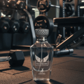 Maison Alhambra Victorioso by Maison Alhambra 100ml EDP Men — A clear glass perfume bottle with a silver cap sits on a gym floor, with dumbbells and weight racks in the blurred background. | Maison Alhambra Victorioso by Maison Alhambra 100ml EDP Men — Botol minyak wangi kaca jernih dengan penutup perak terletak di