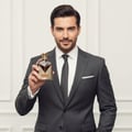 Maison Alhambra Victorioso Heroic by Maison Alhambra 100ml — A man in a dark suit holds a gold and brown patterned perfume bottle in his right hand, looking at the camera. | Maison Alhambra Victorioso Heroic by Maison Alhambra 100ml — Seorang lelaki berbaju gelap memegang botol minyak wangi bermotif emas dan coklat di