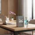 Maison Alhambra Victorioso by Maison Alhambra 100ml EDP Men — A grey perfume box and a blue glass bottle sit on a wooden desk next to an open laptop and a potted orchid. | Maison Alhambra Victorioso by Maison Alhambra 100ml EDP Men — Kotak minyak wangi kelabu dan botol kaca biru diletakkan di atas meja kayu