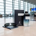 Maison Alhambra Yeah by Maison Alhambra 100ml EDP Men — A bottle of Yeah! Man fragrance and its box are displayed on a counter in an airport terminal, with sunglasses and a passport in the foreground. | Maison Alhambra Yeah by Maison Alhambra 100ml EDP Men — Sebotol pewangi Yeah! Man dan kotaknya dipamerkan di atas