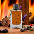 Maison Alhambra Your Touch Intense — A clear glass perfume bottle with amber liquid and a black ribbed cap, set against a backdrop of flames and wood, with lavender sprigs and a pine cone in the foreground. | Botol minyak wangi kaca jernih dengan cecair berwarna ambar dan penutup berjalur hitam, berlatar belakang api