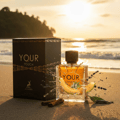 Maison Alhambra Your Touch Man by Maison Alhambra – 100 ml — A perfume bottle and its box sit on a sandy beach at sunset, with a slice of cantaloupe and lavender sprigs around it. | Maison Alhambra Your Touch Man by Maison Alhambra – 100 ml — Botol minyak wangi dan kotaknya diletakkan di pantai berpasir semasa