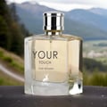 Maison Alhambra Your Touch Women by Maison Alhambra – 100 ml — A clear glass perfume bottle with a silver cap sits on a dark surface, with a blurred mountainous landscape in the background. | Maison Alhambra Your Touch Women by Maison Alhambra – 100 ml — Botol minyak wangi kaca jernih dengan penutup perak diletakkan